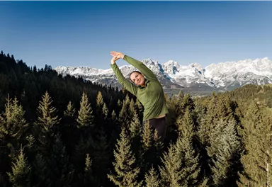 A person is stretching in a wooded landscape with snow-covered mountains in the background. The sky is clear and blue.