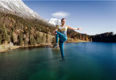 A person is doing a yoga pose over a clear, blue lake. In the background, mountains and trees can be seen.
