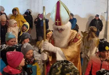 Ein festlicher Anlass mit vielen Kindern, die um eine historische Figur versammelt sind. Der Mann in dem goldenen Gewand hat einen langen Bart und trägt eine Mitra.