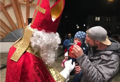 A costumed Santa Claus is speaking with a father and his small child. In the background, other people and a festive atmosphere can be seen.