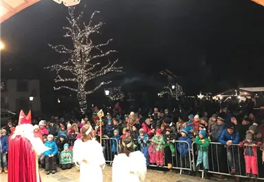 A large crowd gathers at a festive event. In the foreground, figures in costumed garments stand, surrounded by festive lighting.