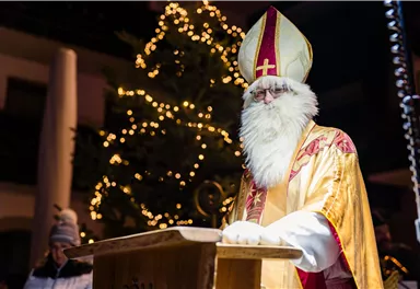 Ein Mann in religiöser Gewandung mit einem weißen Bart steht vor einem Weihnachtsbaum. Es ist eine festliche Atmosphäre mit Lichtern im Hintergrund.