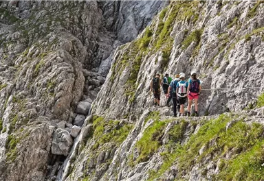 Eine Gruppe von Wanderern geht entlang eines steilen Felsens in den Bergen. Um sie herum sind raues Gestein und grünes Gras.