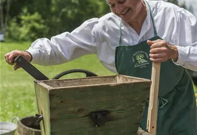 Ein Mann arbeitet an einer alten Saftpresse im Freien. Im Hintergrund sind Bäume und eine grüne Wiese zu sehen.