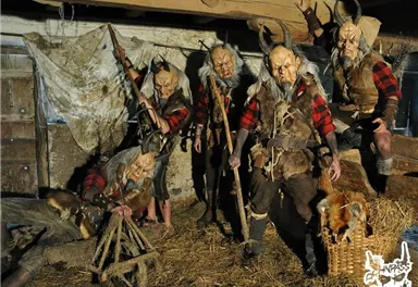 Five masked individuals in traditional clothing stand in a dimly lit room. They are holding wooden swords and appear to be part of a custom or performance.