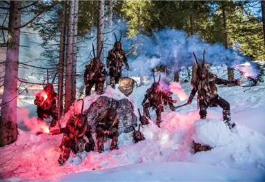 A group of mutated creatures in a snowy forest. They wear terrifying costumes and hold smoke torches in their hands.