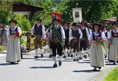 A traditional costume parade with people in festive attire. They happily walk along a street, surrounded by trees and rural scenery.