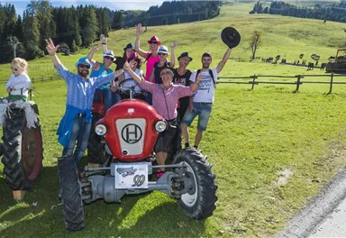 Eine Gruppe von Menschen feiert fröhlich auf einem roten Traktor in einer grünen Wiese. Im Hintergrund sind Berge und Bäume zu sehen.
