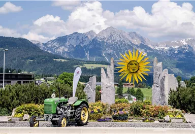 A green tractor stands in front of a sunny, blooming flower bed. In the background, majestic mountains and a bright blue sky can be seen.