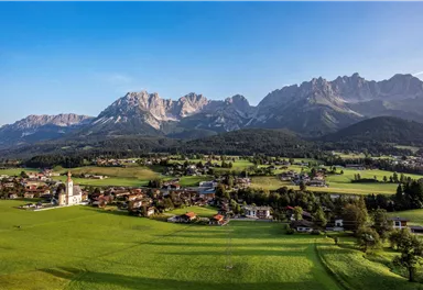 Eine malerische Landschaft mit grünen Wiesen und einem kleinen Dorf. Im Hintergrund erheben sich majestätische Berge unter einem klaren Himmel.