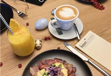 Ein köstliches Frühstück mit Rinderbraten, pochiertem Ei und Kirschtomaten auf einem schwarzen Teller. Im Hintergrund sind eine Tasse Kaffee und ein Glas Orangensaft zu sehen.