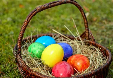 A basket with colorful Easter eggs lies on the grass. The eggs are in various colors like blue, yellow, red, and green.