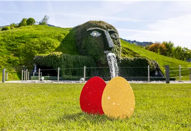 Eine grüne Hügellandschaft mit einer großen, kunstvollen Wasserspeier-Figur im Hintergrund. Im Vordergrund stehen zwei bunte Ostereier, eines rot und das andere gold.