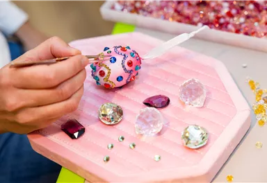 Eine Hand dekoriert ein buntes Ei mit Strasssteinen. Daneben liegen verschiedene glitzernde Steine auf einem rosa Untergrund.