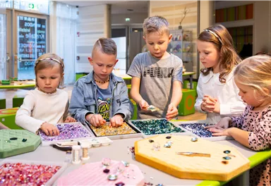 Eine Gruppe von Kindern arbeitet gemeinsam an einem Bastelprojekt. Sie sortieren bunte Perlen und Schmucksteine auf einem Tisch.
