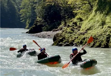 Drei Kajakfahrer paddeln auf einem ruhigen Fluss. Im Hintergrund sind grüne Bäume und eine sonnige Landschaft zu sehen.