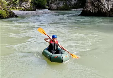 Ein Mensch paddelt in einem kleinen Schlauchboot auf einem ruhigen Fluss. Umgeben von üppiger Natur und hohen Felsen.