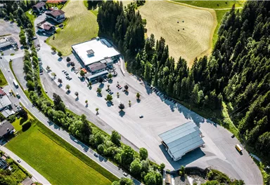 A large parking area with several cars and a small sales booth. Surrounded by green forests and gentle hills.