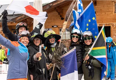 A group of cheerful skiers stands together holding various flags. They are wearing winter sports clothing and smiling at the camera.