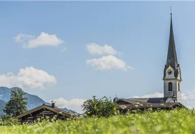 A picturesque landscape with a church and a traditional building in the foreground. The sky is blue with some clouds and the mountains are visible in the background.