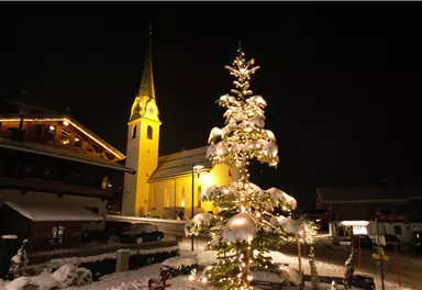A festively decorated Christmas tree stands in the snow, illuminated by warm light. In the background, there is a church with a tall tower, also bathed in warm light.
