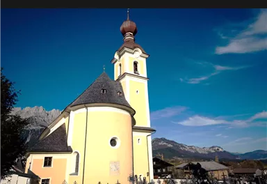 A beautiful church with a yellow facade and a distinctive tower. In the background, mountains and a blue sky can be seen.