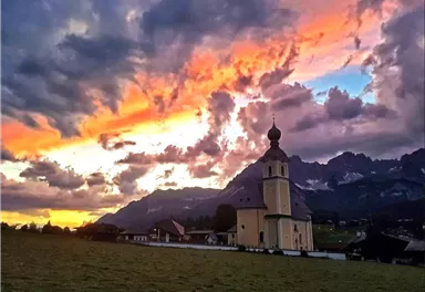 A picturesque church against a spectacular evening backdrop. The sky is filled with vibrant colors and clouds, while the mountains are visible in the background.