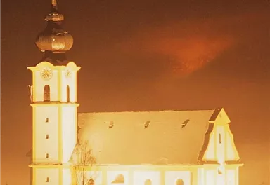 Eine beleuchtete Kirche im Schnee bei Nacht. Der Himmel ist dunkel und der Turm ist gut sichtbar.