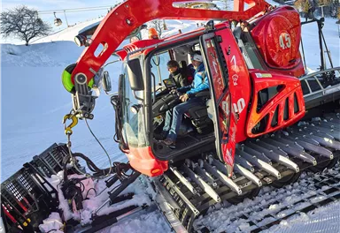 Ein großer roter Pistenbully steht im Schnee. Zwei Personen sitzen im Fahrerhaus und schauen konzentriert.