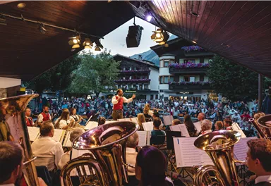 Ein Open-Air-Konzert mit einer Musikkapelle, die auf einer Bühne spielt. Im Hintergrund sind viele Zuschauer und traditionelle Gebäude zu sehen.