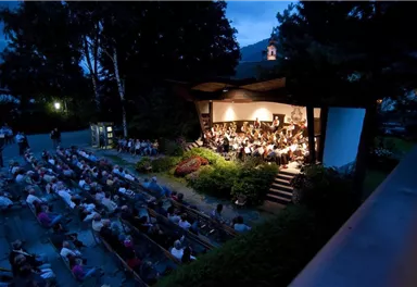 An outdoor concert with an orchestra on a stage. Many spectators are sitting on benches and enjoying the music in a cozy evening atmosphere.