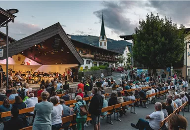 Ein freiluft Konzert mit vielen Zuschauern in einem malerischen Dorf. Im Hintergrund ist ein Kirchturm und die umliegende Landschaft zu sehen.