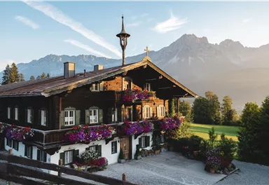 Ein traditionelles, mit Blumen geschmücktes Holzhaus in den Bergen. Im Hintergrund sind majestätische Berge und ein klarer Himmel zu sehen.