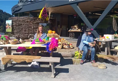 Eine bunte Marktszene im Freien mit fröhlichen Menschen und verschiedenen Waren. Auf den Tischen sind Lebensmittel und Dekorationen aufgestellt.