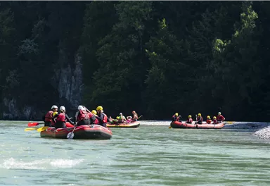 A group of people is boating on a river in inflatable rafts. The surroundings are green and forested.