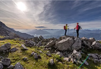 Two hikers are standing on a rock in the mountains, enjoying the view. The sky is clear with soft clouds, and the landscape is green and mountainous.