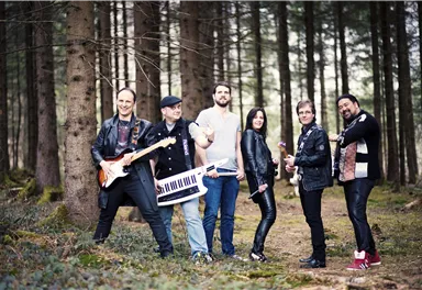 A band stands in the woods and poses with their instruments. The members wear casual clothing and look ready to play music.