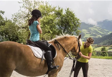 A girl is sitting on a horse and wearing a helmet. A second person is taking care of the horse and stands next to it.