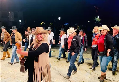 A joyful group dance event in cowboy style outdoors. The participants wear hats and celebrate to the music of a singer with a guitar.
