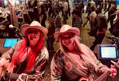 Two women in hats are playing music on a stage. In the background, people dressed in cowboy outfits are dancing in a festive atmosphere.