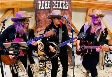 Three female musicians in cowboy hats are playing on stage. In the background, there is a banner with the inscription "ROAD CHICKS".