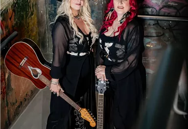 Zwei Musikerinnen stehen in einer Treppe mit Graffiti, eine mit einer akustischen Gitarre und die andere mit einem Banjo. Beide tragen Cowboyhüte und auffällige Outfits.