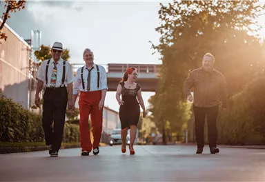 Vier Menschen gehen gemeinsam auf einer Straße, umgeben von Bäumen und Gebäuden. Die Sonne scheint und schafft eine warme Atmosphäre.