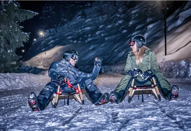Zwei Personen sitzen im Schnee auf Rodeln und lachen sich an. Die Umgebung ist nachts und mit Schnee bedeckt, umgeben von schneebedeckten Bäumen.