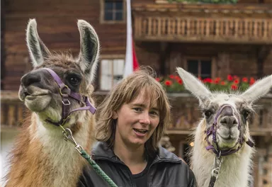 Eine Frau steht mit zwei Lamas vor einem Holzhaus. Im Hintergrund sind Blumen und eine Fahne zu sehen.