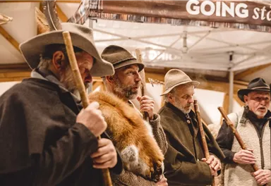 Ein Gruppe von Männern in traditionellen Kleidern steht zusammen und hält Stöcke in der Hand. Sie scheinen an einem kulturellen oder historischen Event teilzunehmen.