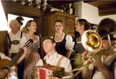 Eine fröhliche Gruppe von Menschen in traditioneller Tracht singt und spielt Musik in einem rustikalen Raum. Es gibt Instrumente wie Gitarre und Trompete, die zur festlichen Stimmung beitragen.