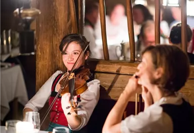 Eine Musikerin spielt Geige in einem gemütlichen Restaurant. Im Hintergrund sind weitere Gäste zu sehen.