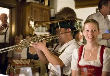 Eine festliche Atmosphäre in einem traditionellen Restaurant. Musiker spielen auf Instrumenten, während Gäste in Trachten lächeln und die Unterhaltung genießen.