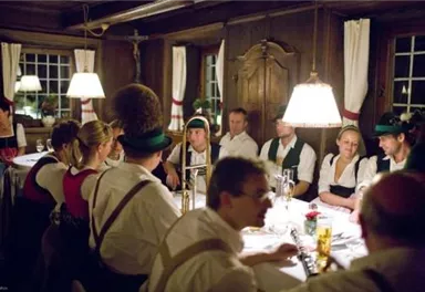 Eine gesellige Runde von Gästen in einem traditionellen Restaurant. Die Atmosphäre ist gemütlich, mit warmem Licht und bayerischer Tracht.